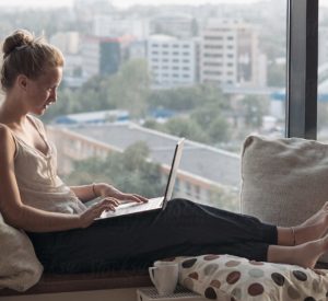 woman lounges while working on laptop near large window
